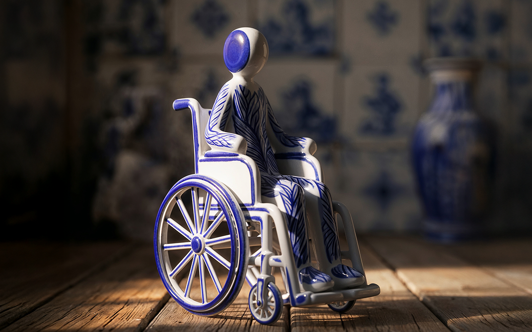Blue-and-white ceramic figurine seated in a wheelchair on a wooden surface, with a blurred blue and white tile background.